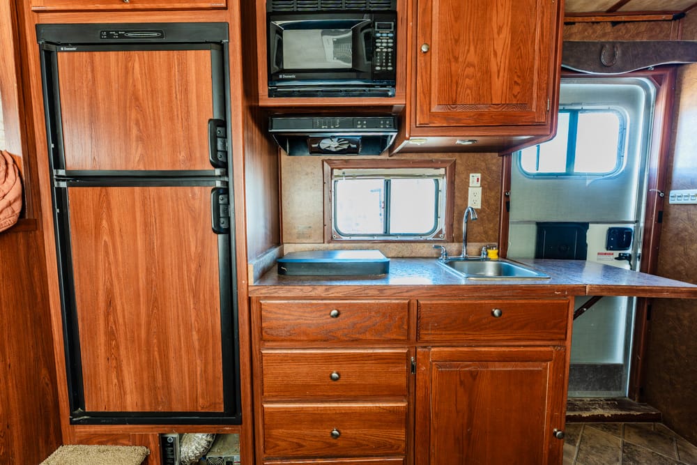 Wooden RV kitchen featuring a tall double-door fridge on the left, microwave above, and a sink with a window over the counter.