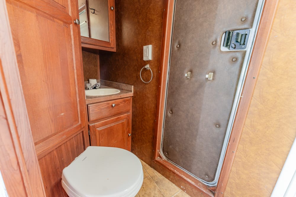 Compact RV bathroom with wooden cabinetry, sink, and toilet, plus a brown shower door enclosure nearby