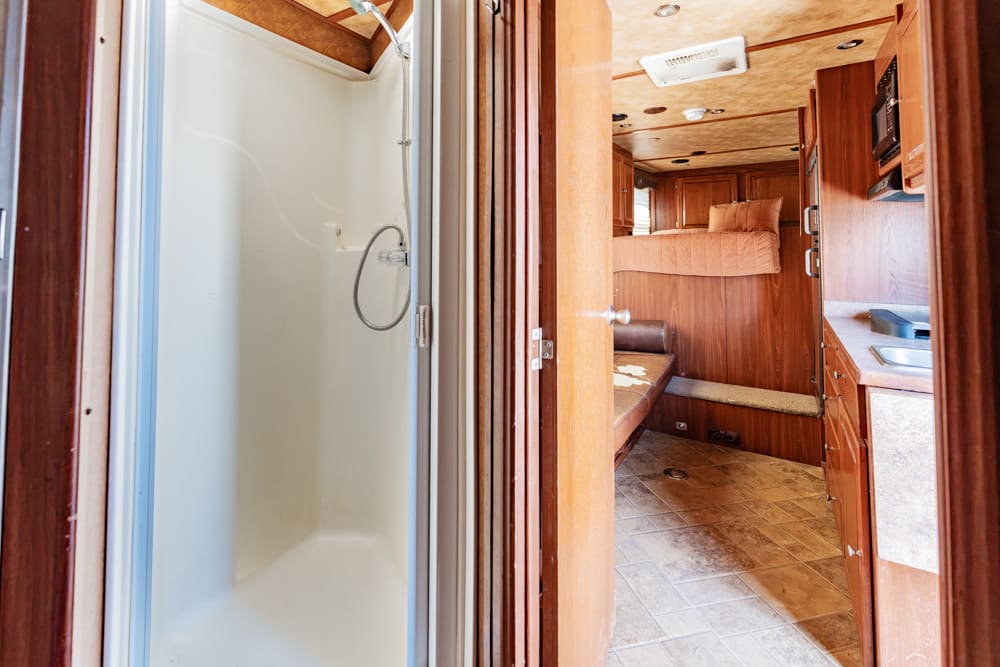 Interior of a wood-paneled RV with a white shower stall to the left and a kitchen area to the right, leading to a bedroom in the back.