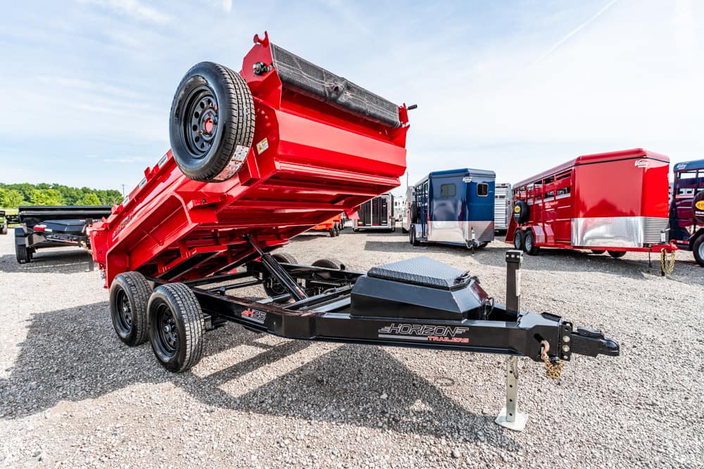 Red dump trailer raised at an angle on a black Horizon trailer frame in a gravel lot, with other trailers in the background.