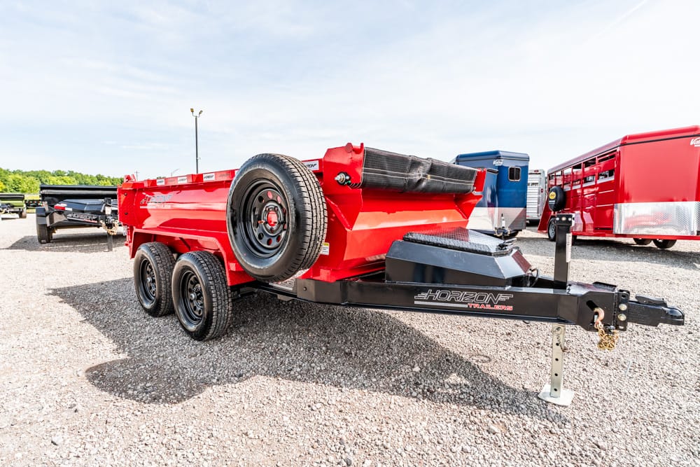 Red Horizon tilt trailer with a spare tire mounted on the side, parked on a gravel lot with other trailers nearby.