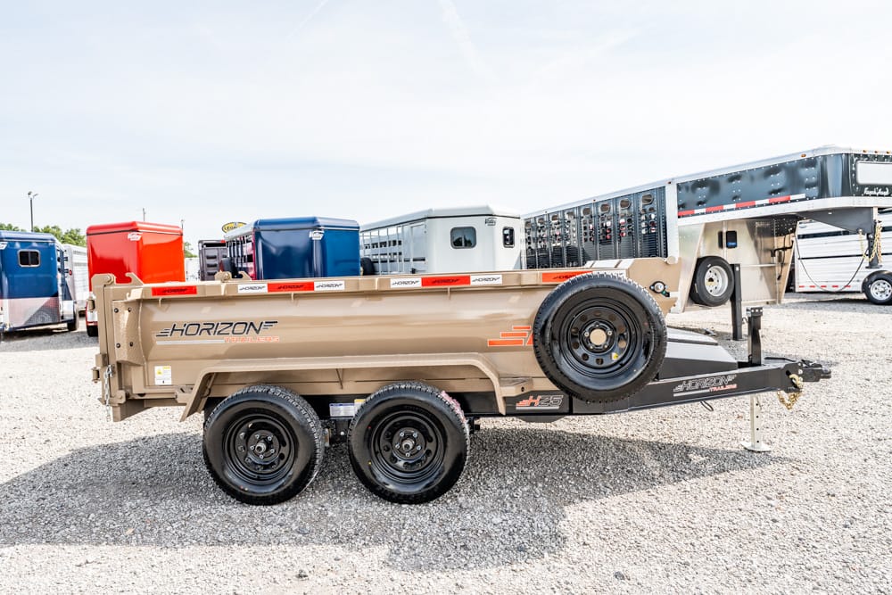 Beige Horizon utility trailer with a spare tire mounted on the front, two axles, parked on a gravel lot with horse trailers and portable toilets in background