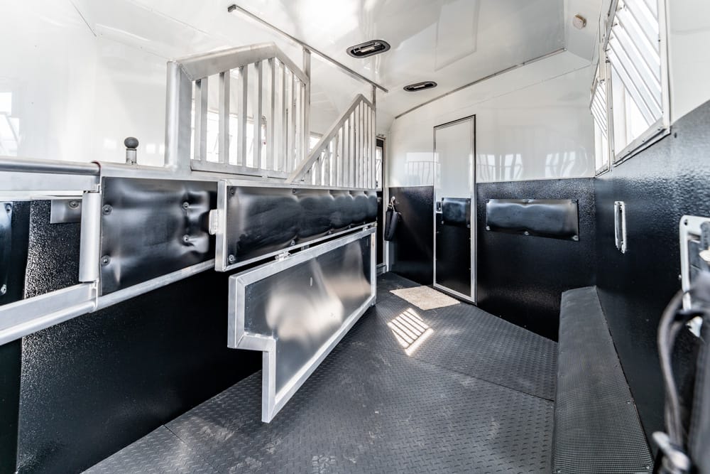Interior of a black-and-silver horse trailer showing padded stalls, metal dividers, and a diamond-plate floor for secure animal transport, daylight from windows at the rear.
