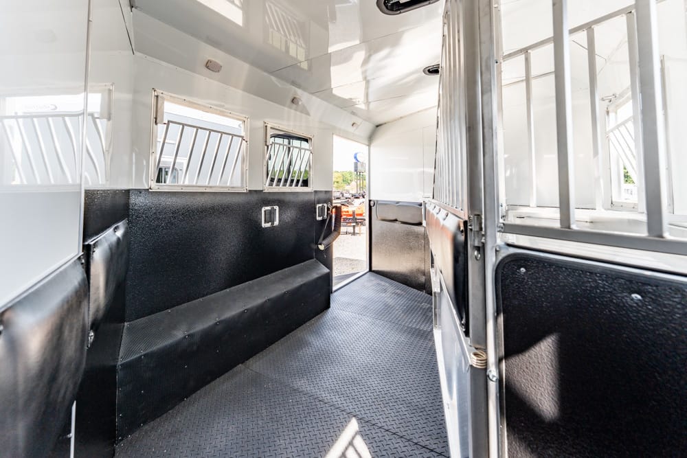 Interior of a white horse trailer with black padded walls, diamond-plate floor, and barred windows along the side corridors.