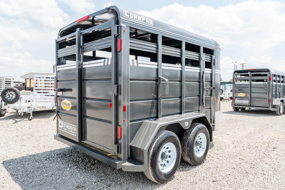 Dark gray CornPro livestock trailer with tandem axles on a gravel lot; rear doors open and side vents visible.