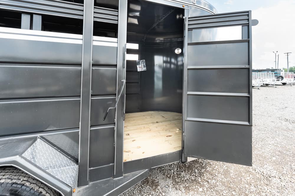 Rear of a black metal trailer with its double doors open, showing a wooden floor inside and a gravel lot outside.