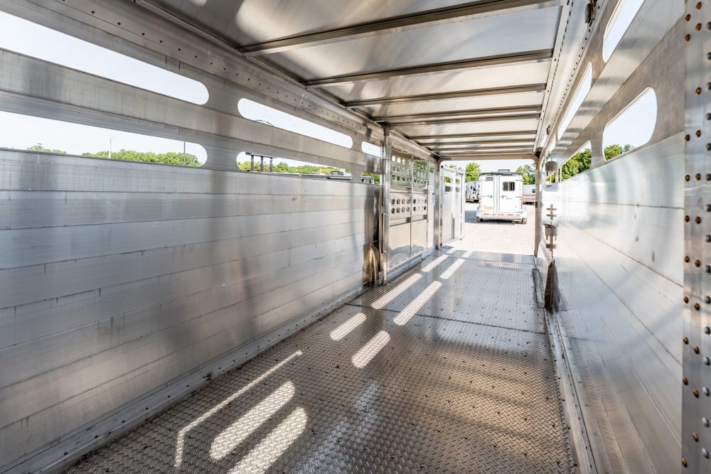 Interior of an empty aluminum livestock trailer with a diamond-plate floor and long cargo space, rear door open to a sunlit yard.