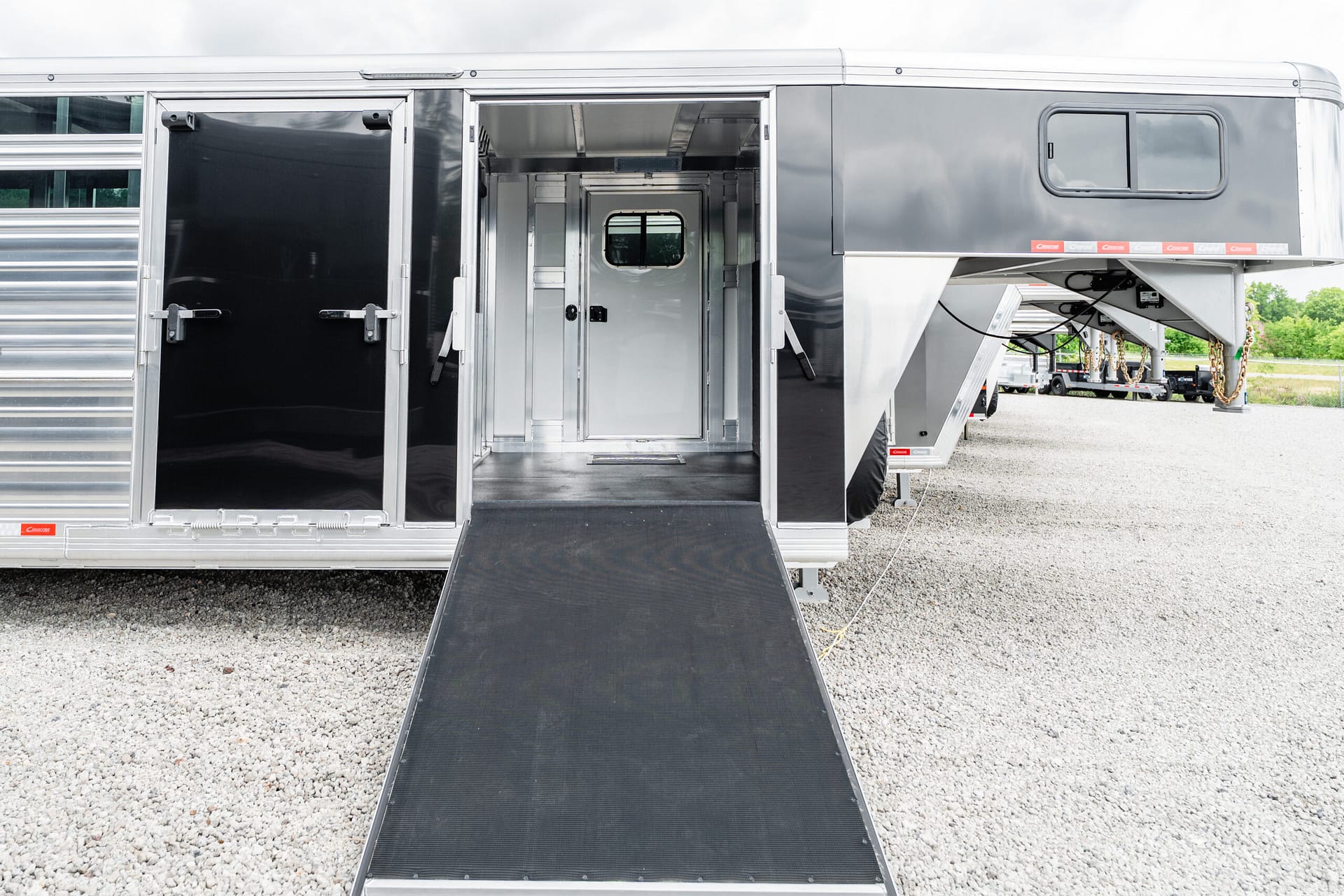 Silver horse trailer with its loading ramp extended, doors open to reveal the interior stalls on a gravel lot.] ,