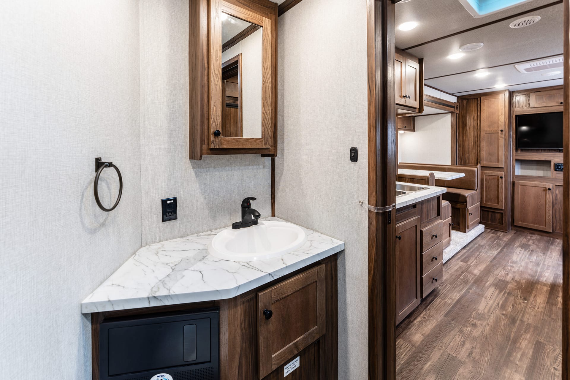 RV bathroom interior with marble countertop sink, wooden cabinet, mirror cabinet, towel ring, and beige walls leading to a living area.