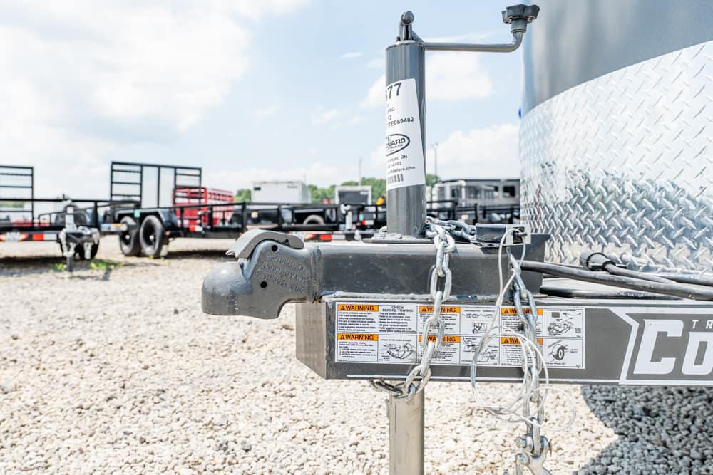 Close-up of a trailer hitch with safety chains and warning labels on a gravel lot in the background of other trailers.