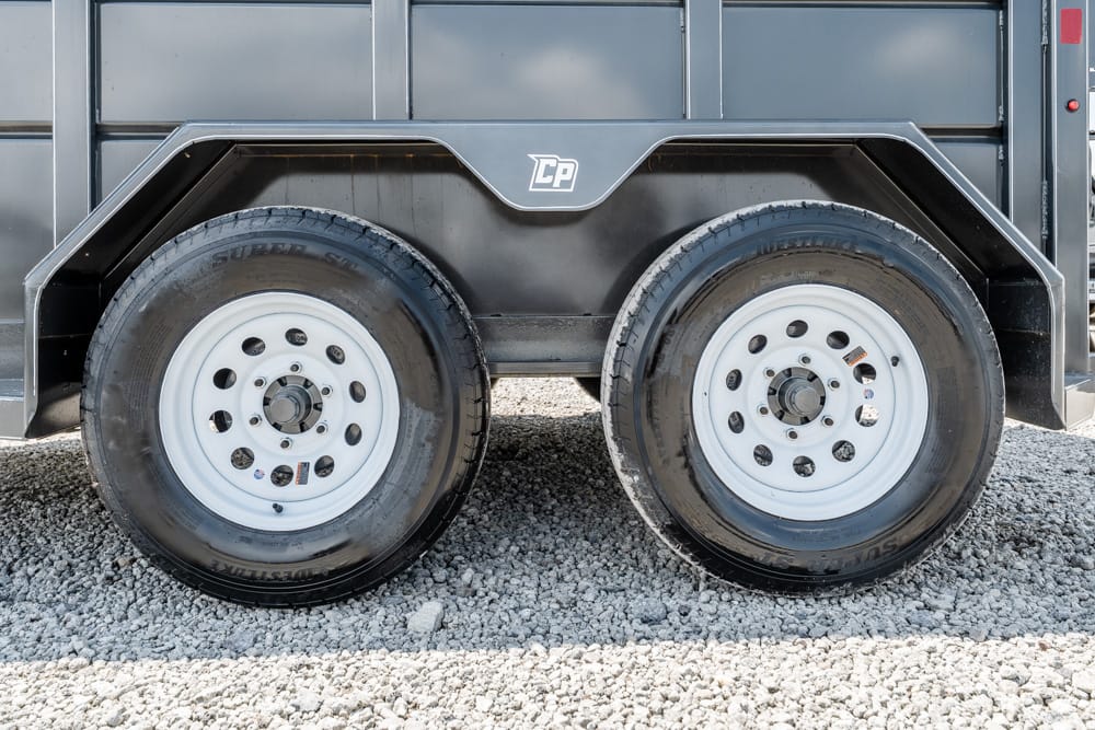Twin trailer wheels with white rims on a black utility/dump trailer parked on gravel, showing the tandem axle setup.