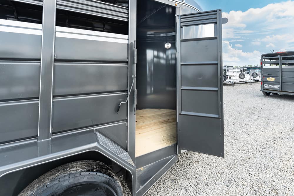 Open horse trailer showing wooden floor inside, with other trailers in a gravel lot outside.