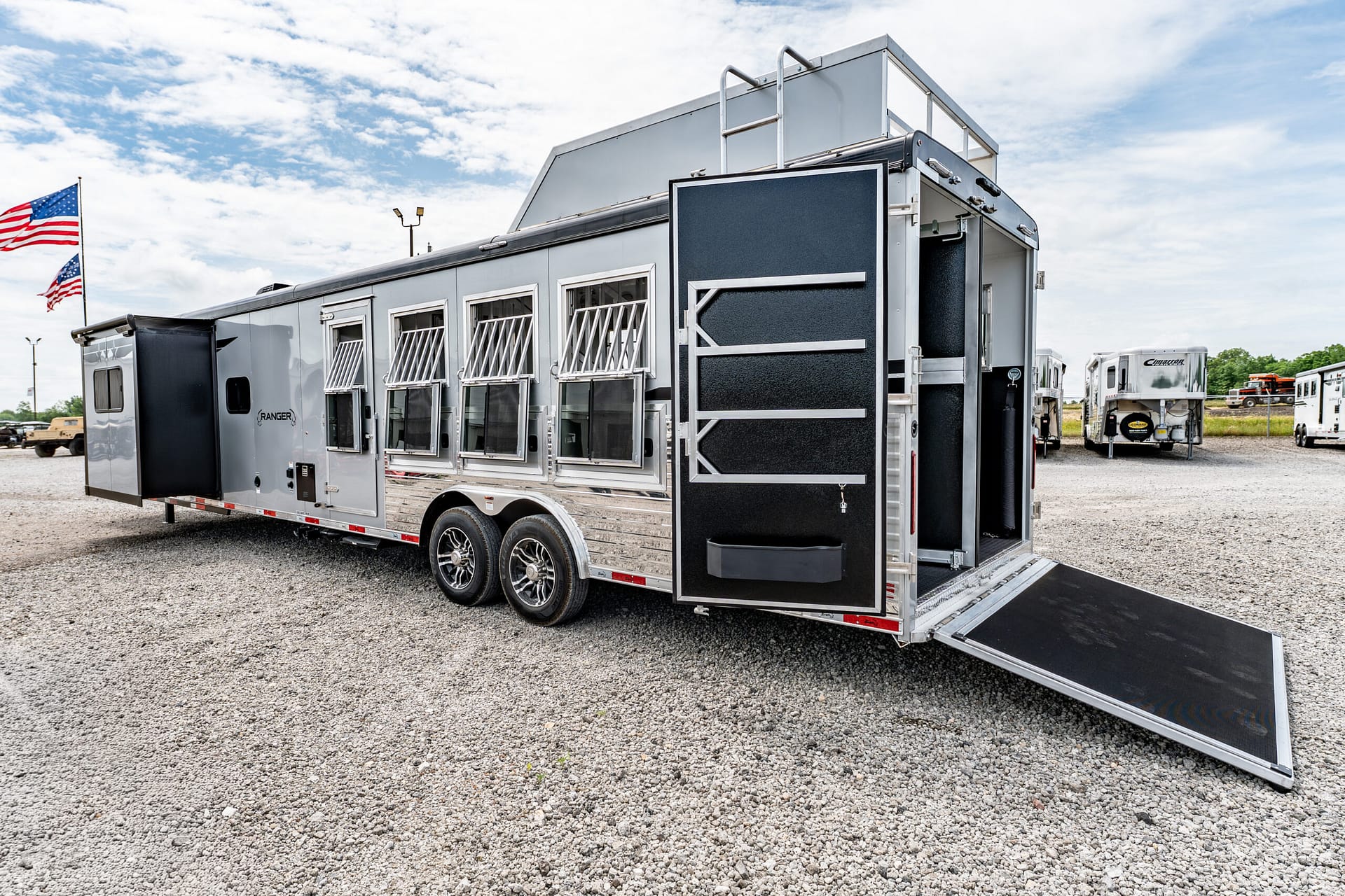 Large silver horse trailer with an open rear ramp on a gravel lot; American flags in the background under a cloudy sky