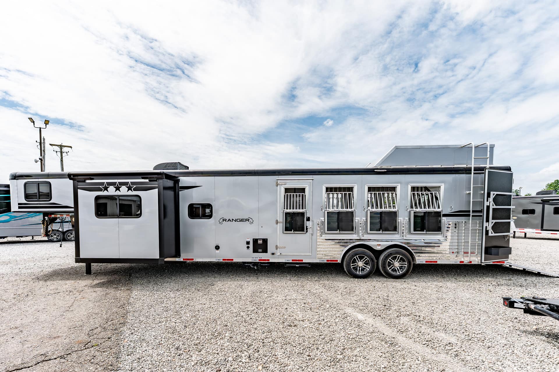 Long silver travel trailer with multiple windows and a curved black awning, parked on gravel under a blue, cloudy sky.