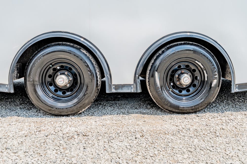 Pair of black trailer wheels on a gravel surface, side-by-side under a light gray trailer body for stability and support.