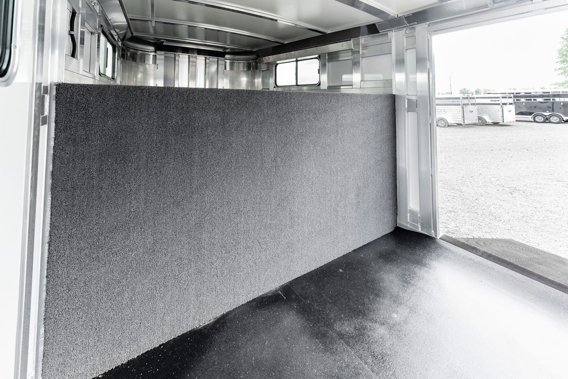 Interior of a metal horse trailer with a tall dark gray carpeted divider and aluminum walls, seen from the back door overlooking other trailers outside