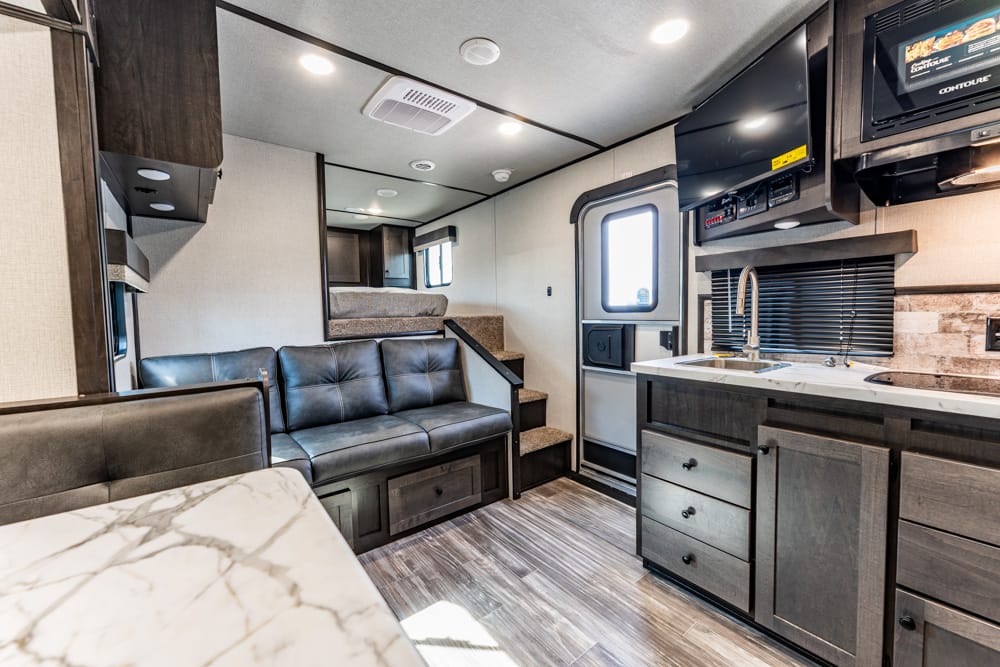 Interior of a modern RV with a gray leather couch, marble countertop, and a kitchen area including sink, stove, and microwave; stairs lead to a loft bunk area on the left.