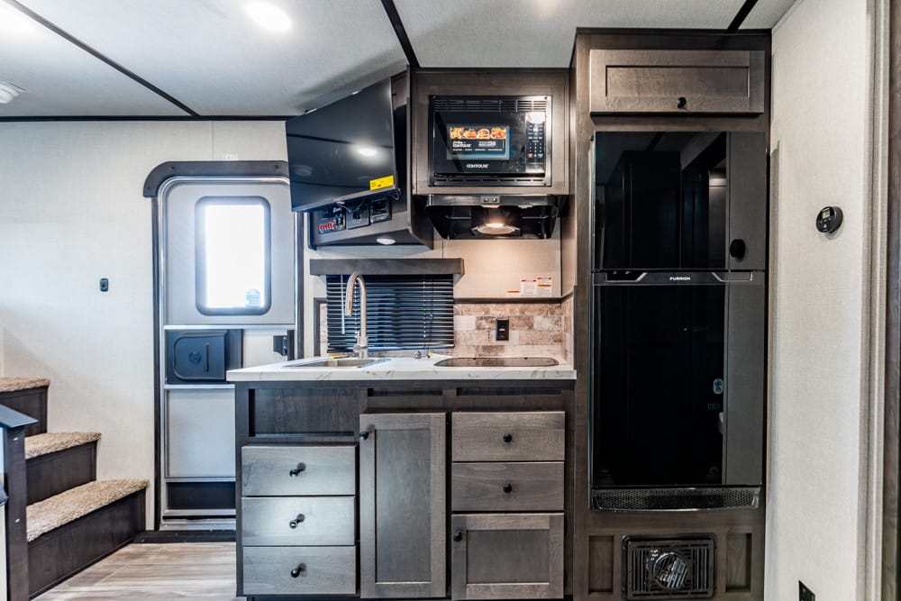 Interior of an RV kitchen with dark wood cabinetry, stainless sink, stove, microwave, and a tall refrigerator.