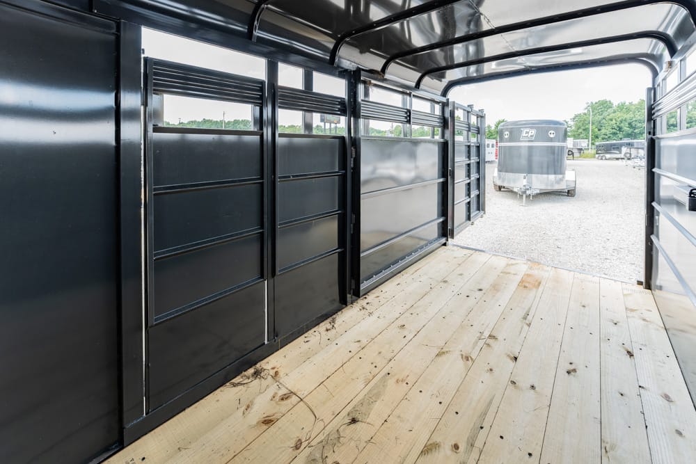Interior of a black horse trailer with partitioned stalls and a light wooden floor, opening to a gravel lot with another trailer in view.