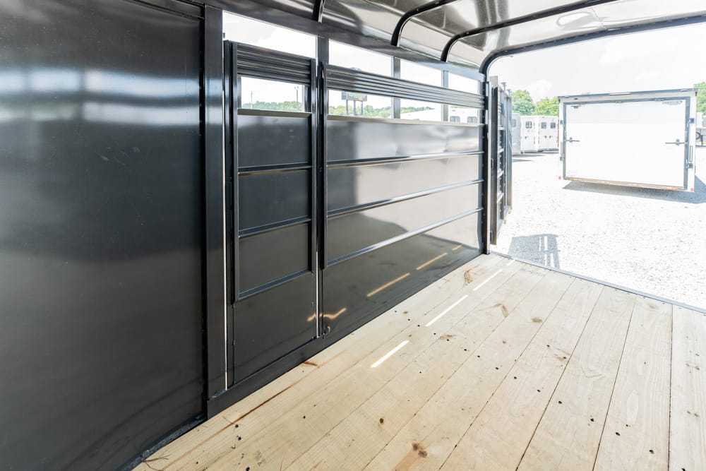 Interior of a black metal trailer with a wooden floor and an open side gate, parked among other trailers on a gravel lot.