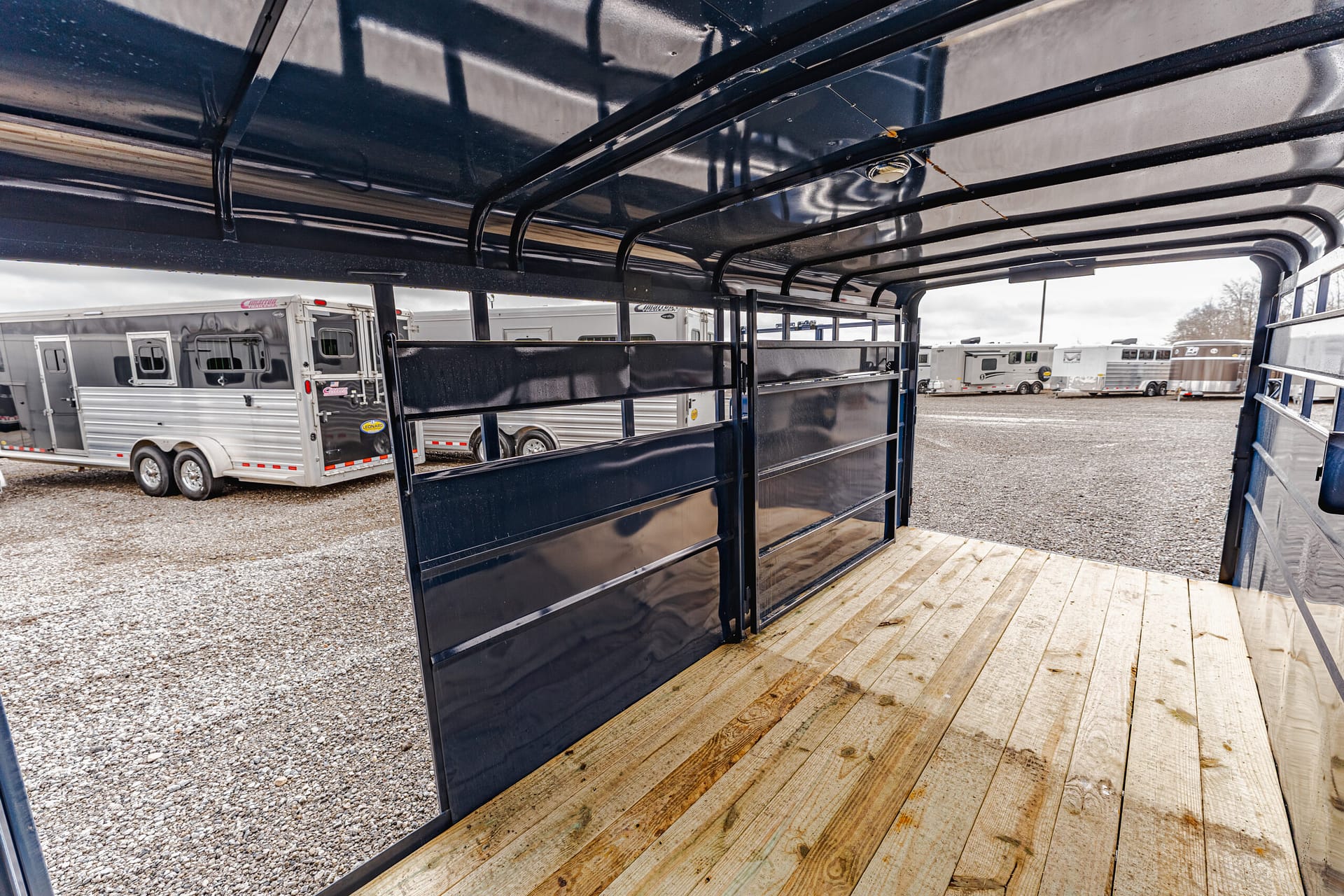 Interior of a black horse trailer with metal partitions and a wooden floor, parked among other trailers on a gravel lot beside a cloudy sky.