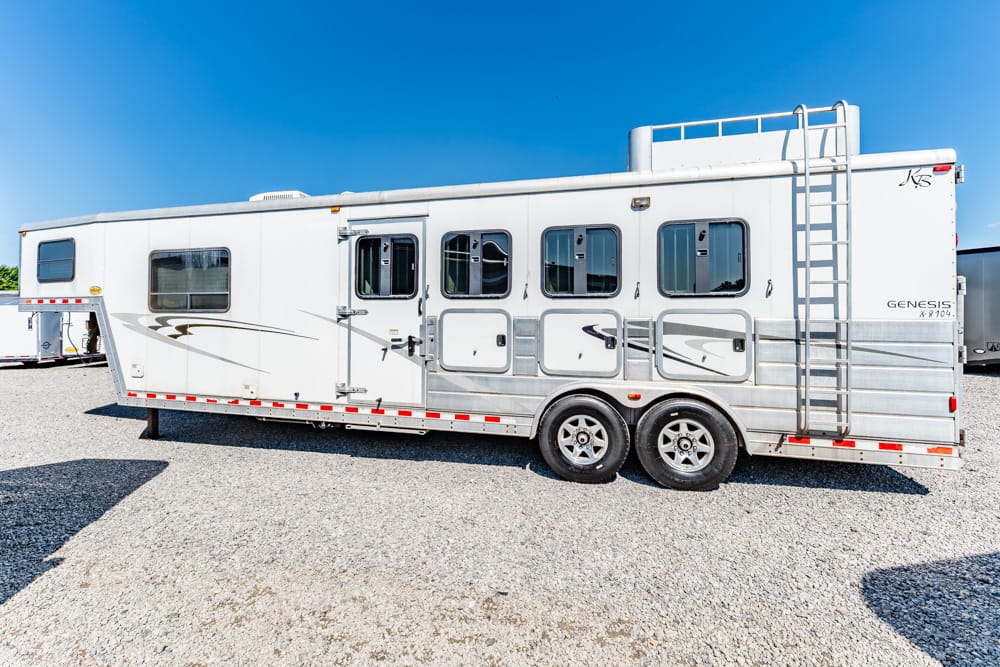 White Genesis horse trailer with multiple windows and a rear ladder, parked on gravel under a clear blue sky