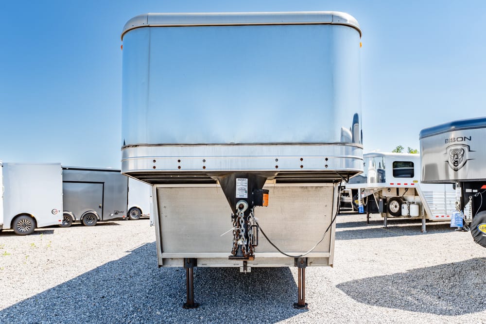 Front view of a large silver trailer on a gravel lot, with a hitch and chains in the foreground and other trailers in the background under a clear blue sky.