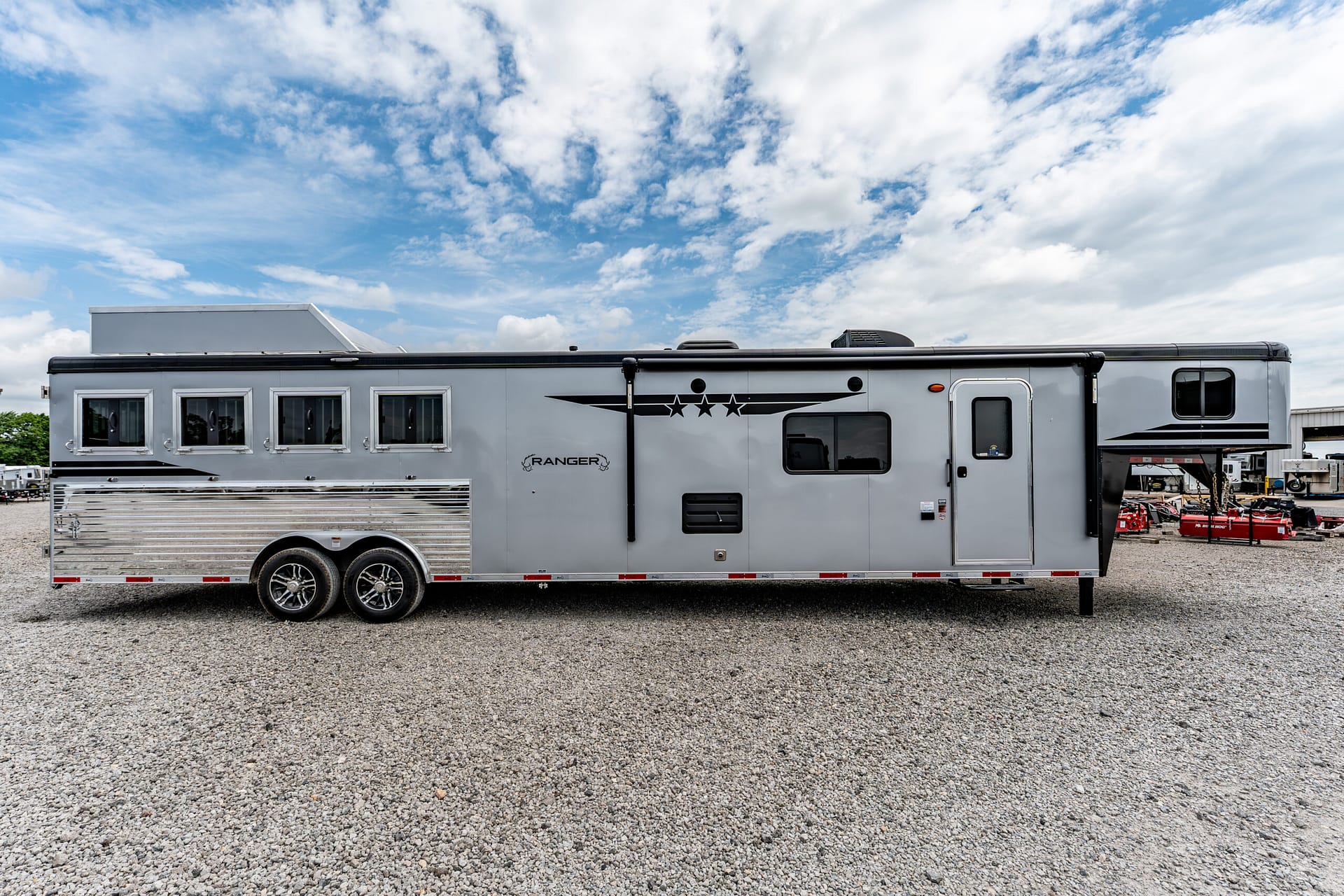 Long silver travel trailer with three windows and decorative stars, parked on a gravel lot under a blue sky with clouds.
