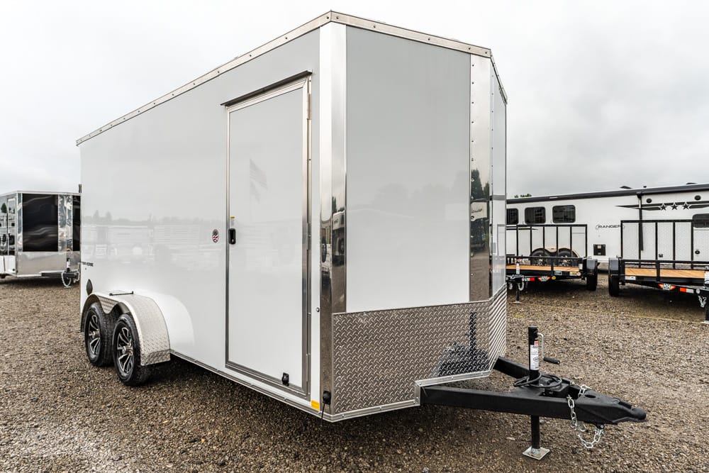 White enclosed cargo trailer with two axles and diamond-plate front on a gravel lot.