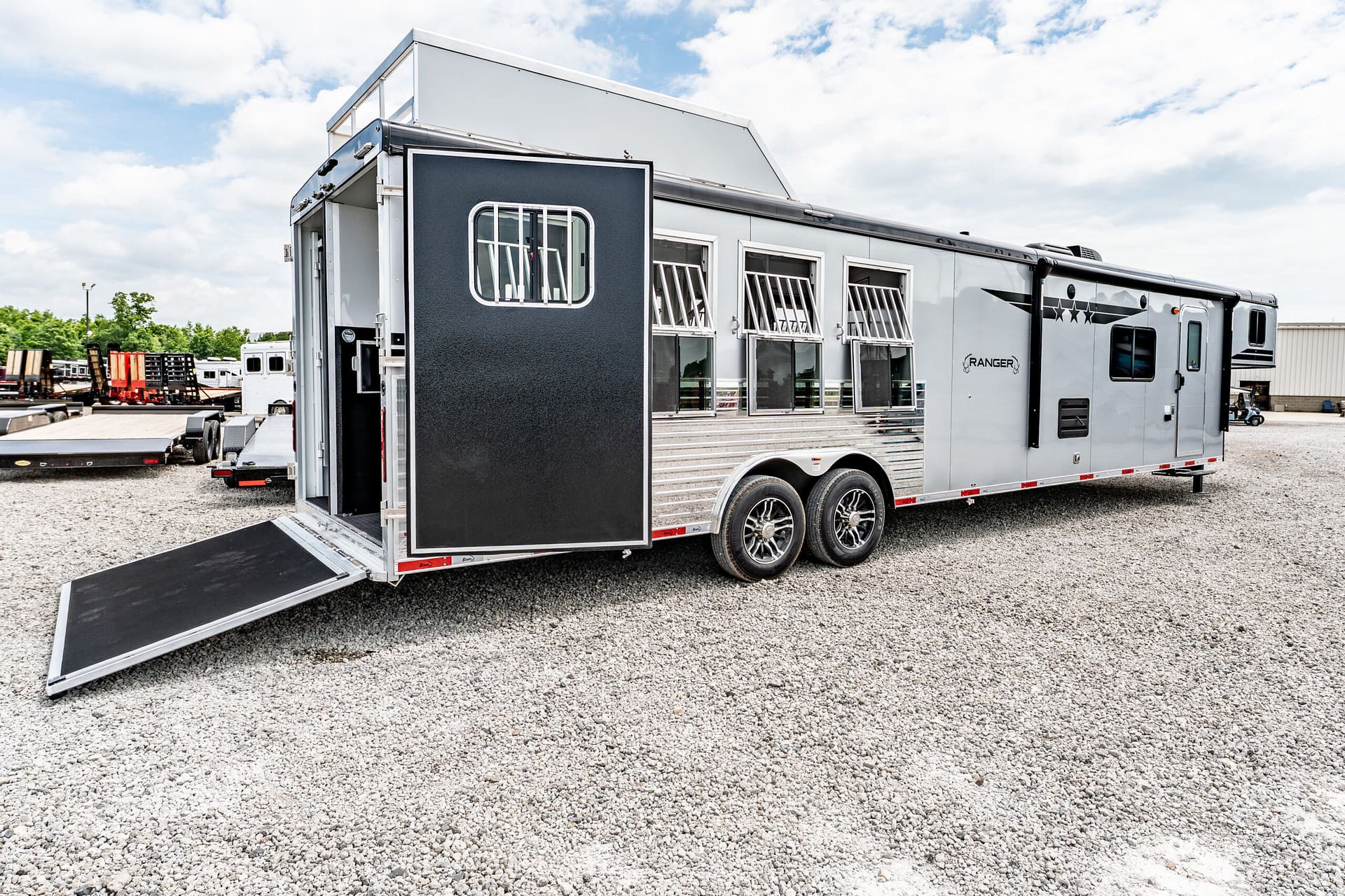 Large silver horse trailer with a lowered ramp, parked on gravel in a lot, windows with bars along the side.
