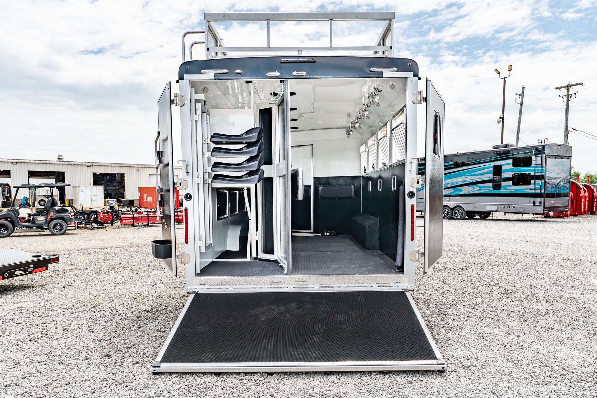 Open back of a metallic trailer with folded black seats inside, a ramp lowered to gravel ground, and other vehicles in the background.