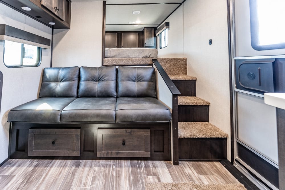 Living area in an RV with a dark leather sofa, built-in under-seat storage drawers, and carpeted stairs to a loft bed above on the right side.