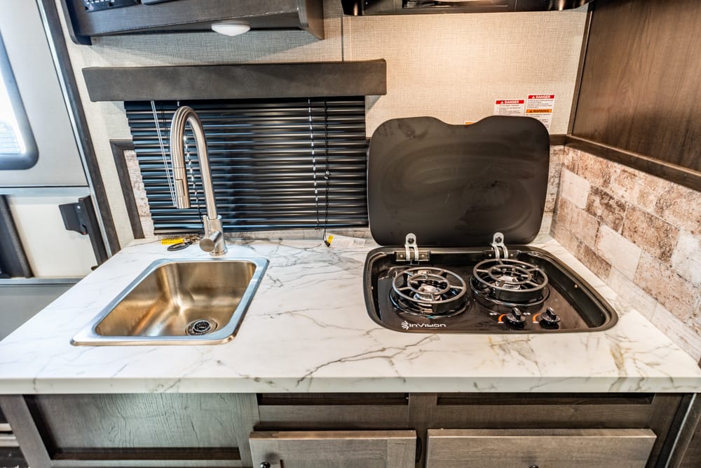 RV kitchen: stainless steel sink and two-burner gas stove on a marble countertop with lid open and window blind in the background