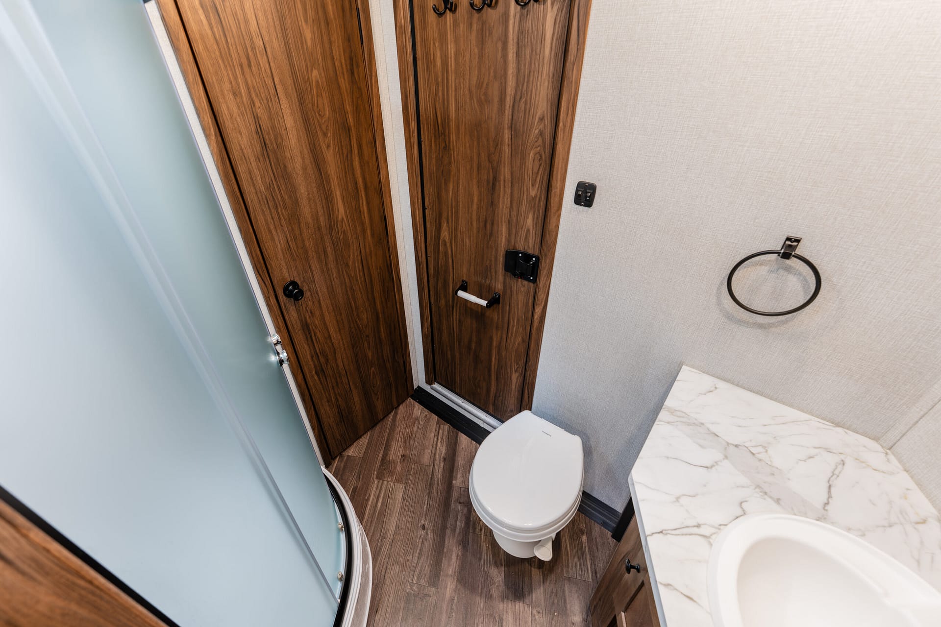 Compact bathroom with curved glass shower, wood grain closet doors, white toilet, and marble countertop in a small space.