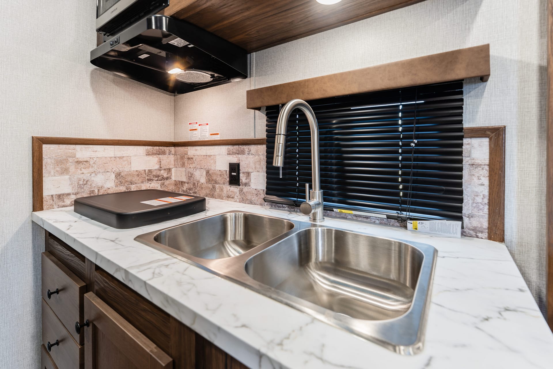 RV kitchenette: stainless double sink, gooseneck faucet, marble countertop, dark wood cabinets, and a window with black blinds.