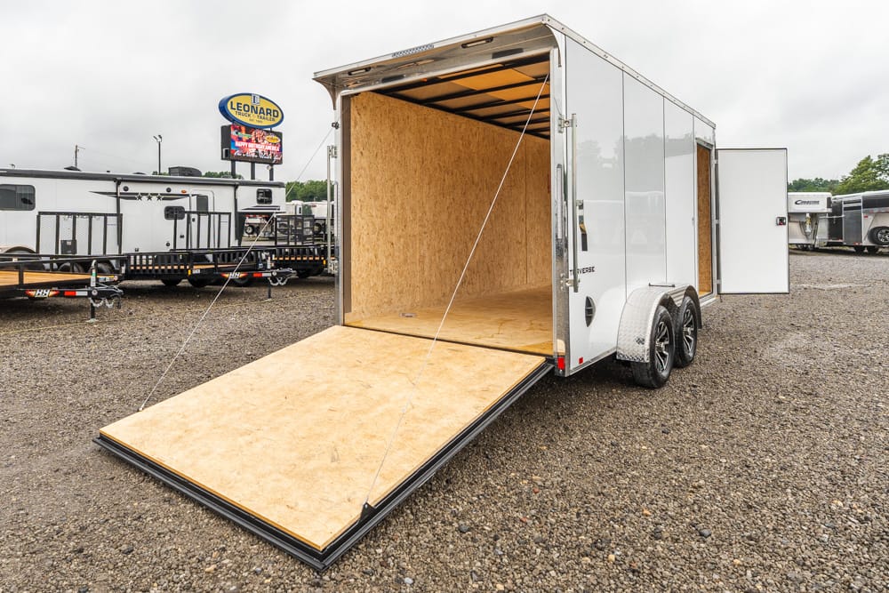 Open cargo trailer with a wooden ramp lowered to the ground on a gravel lot, ready for loading.