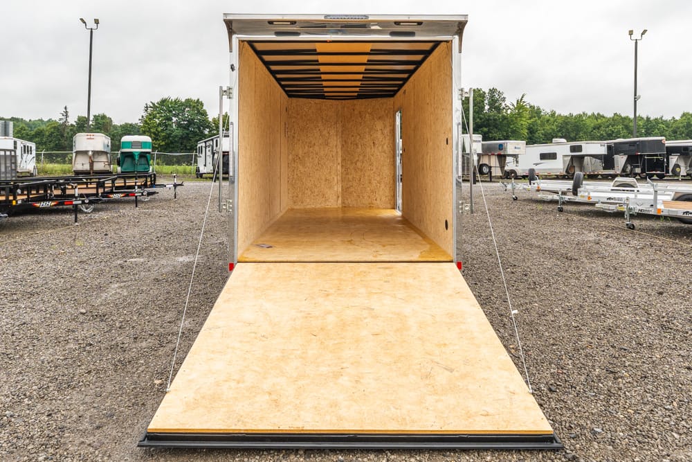 Open metal trailer with a plywood floor and walls, ramp down for loading, in a gravel lot with other trailers nearby.