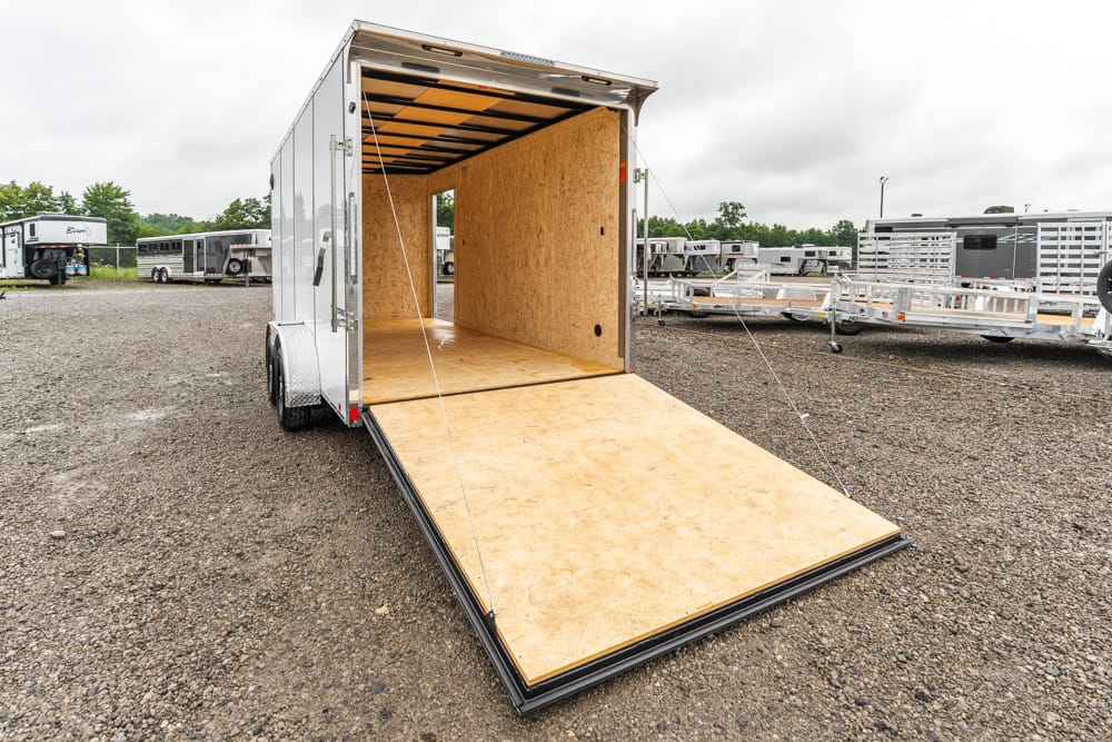 Open cargo trailer with ramp lowered on a gravel lot, showing plywood interior and tie-down ropes nearby.