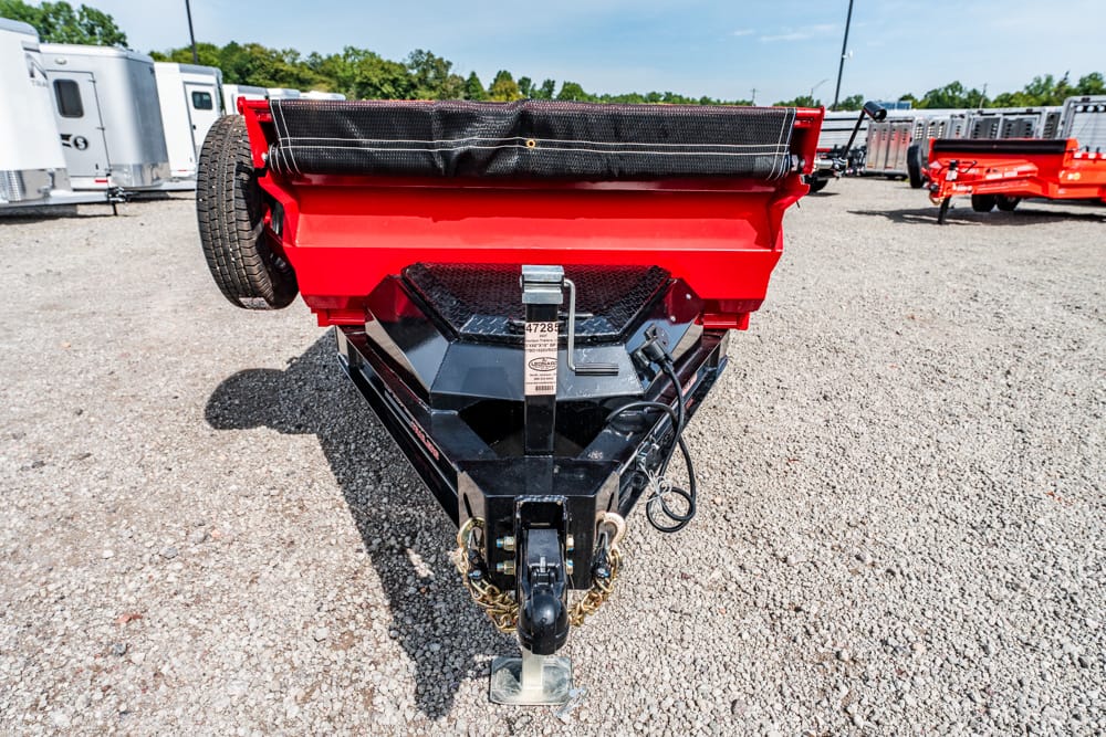 Front view of a red dump trailer hitch with a black frame and safety chains on a gravel lot, with silver trailers in the background.