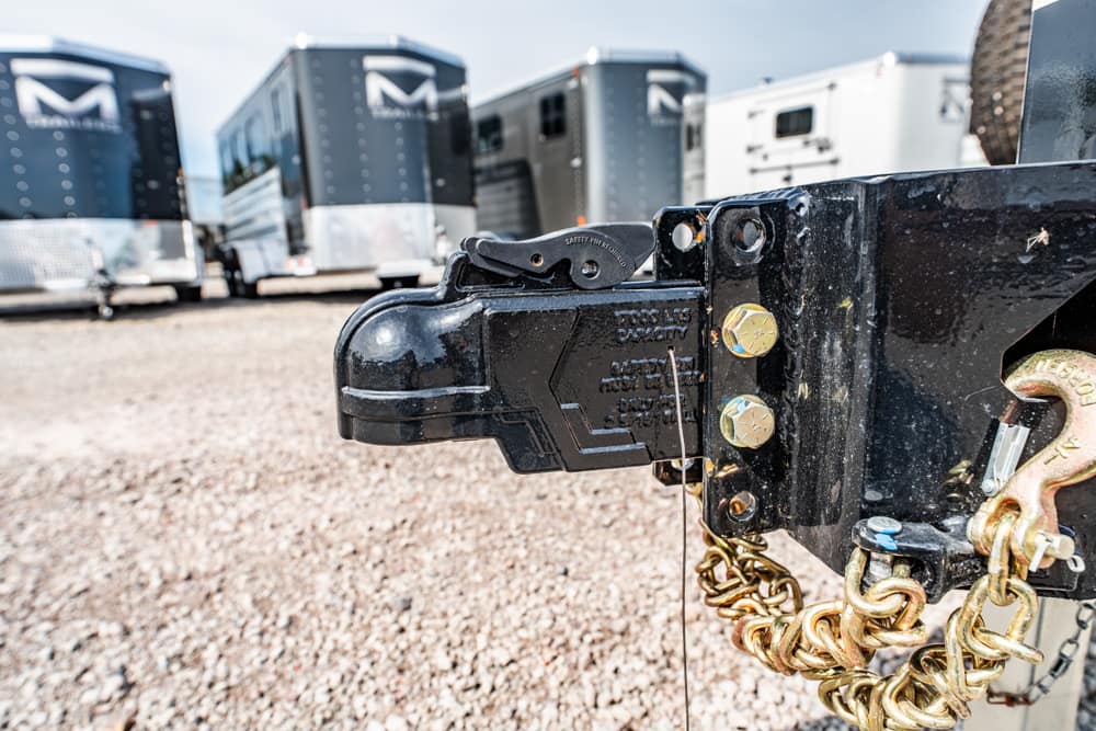 Close-up of a black trailer coupler with a safety latch and gold chains, attached to a trailer in a gravel lot with other trailers in the background.