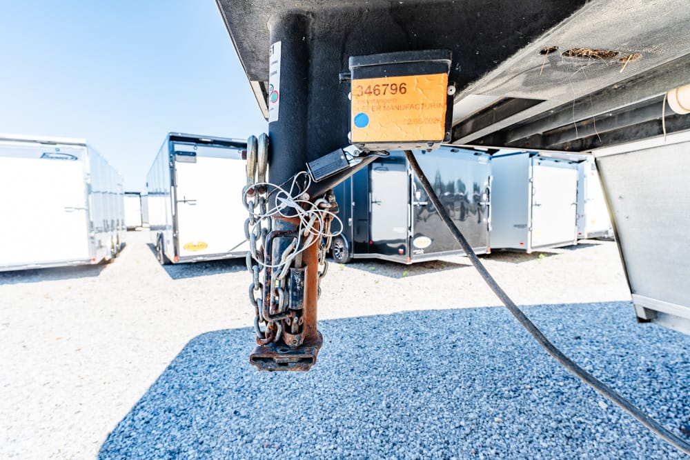 Close-up of a rusty trailer hitch with tangled chains and an orange tag, under a black trailer in a lot of parked trailers.