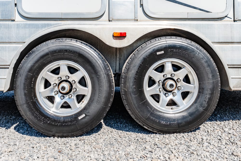 Two rear trailer wheels with black tires and silver rims on a gravel surface.