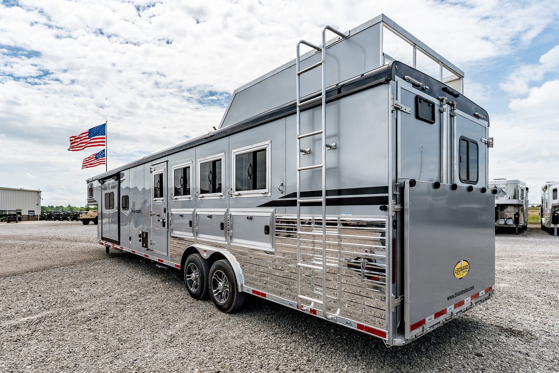 Large silver utility trailer with a metal ladder on the side, parked on a gravel lot under a partly cloudy sky with American flags in the background.