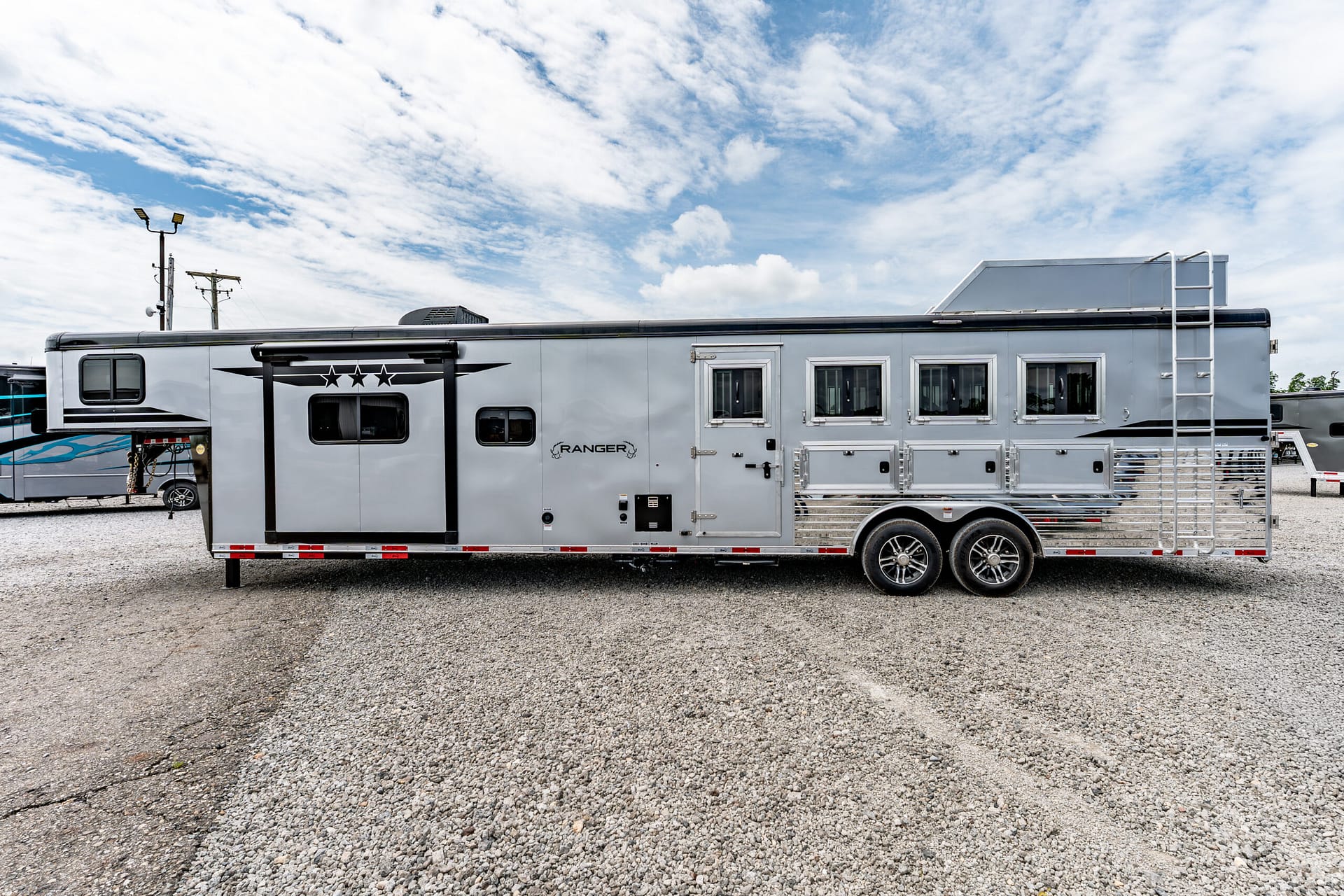 Long silver horse trailer parked on a gravel lot with a rear ladder and a blue sky above.
