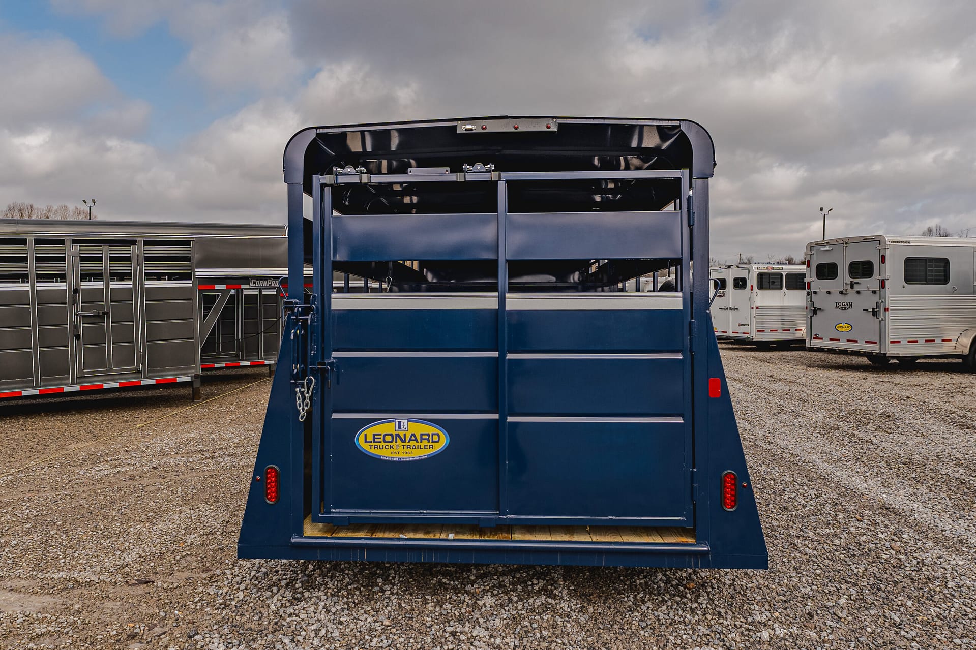 Rear view of a blue trailer with an open top and Leonard Truck & Trailer logo, parked on a gravel lot with other trailers in the background.