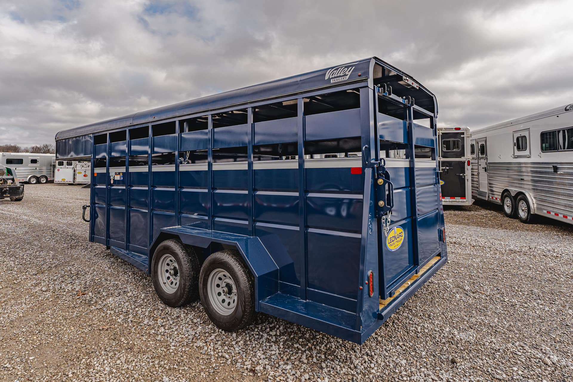 Blue livestock trailer with open side slats parked on a gravel lot, other trailers visible in the background under a cloudy sky.