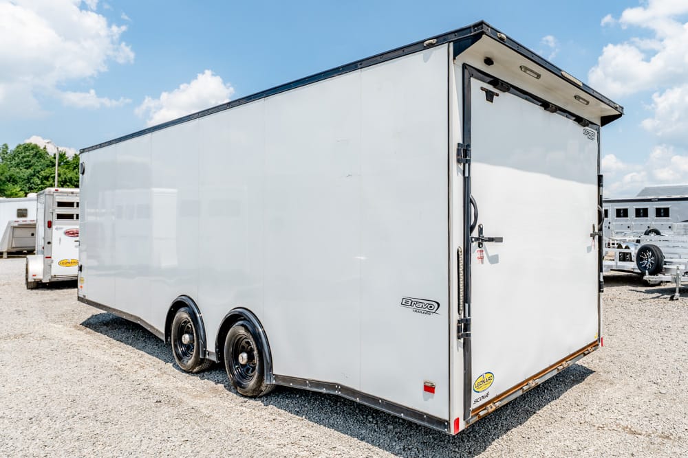 White enclosed cargo trailer with dual axles parked on a gravel lot, rear doors closed and other trailers in the background.