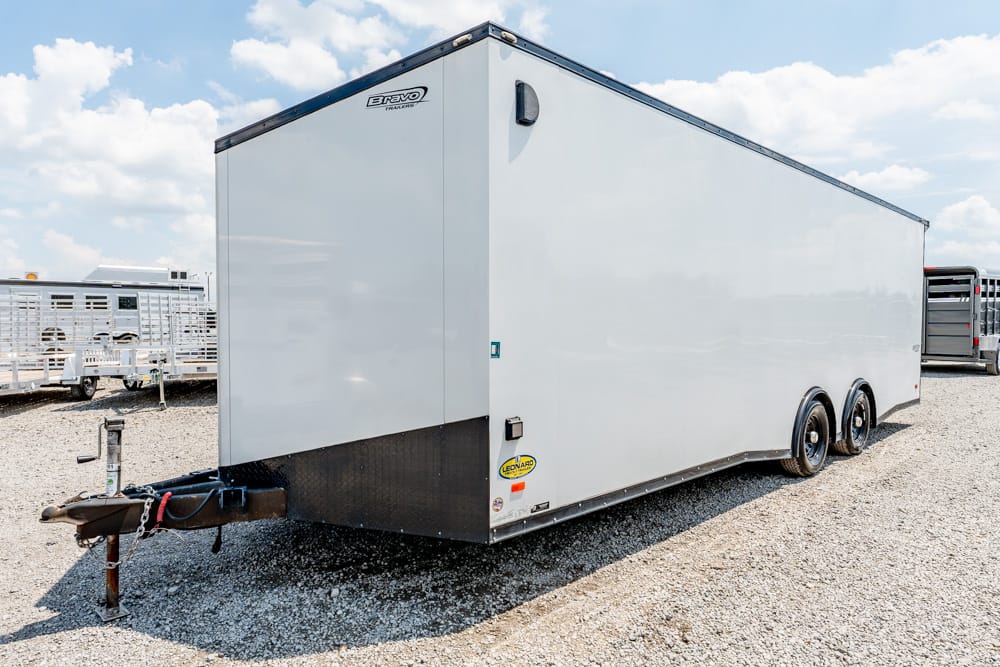 White enclosed cargo trailer with dual axles parked on a gravel lot, front hitch visible in foreground
