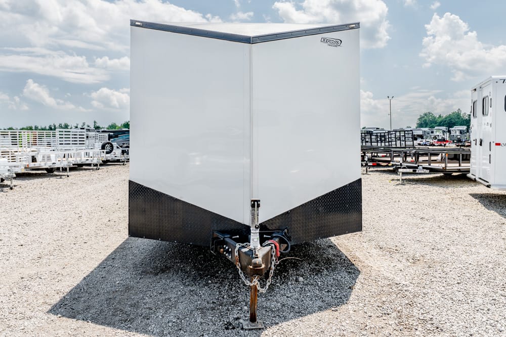 Front view of a white enclosed cargo trailer with black diamond-plate lower front, hitch and safety chains visible in a gravel lot.