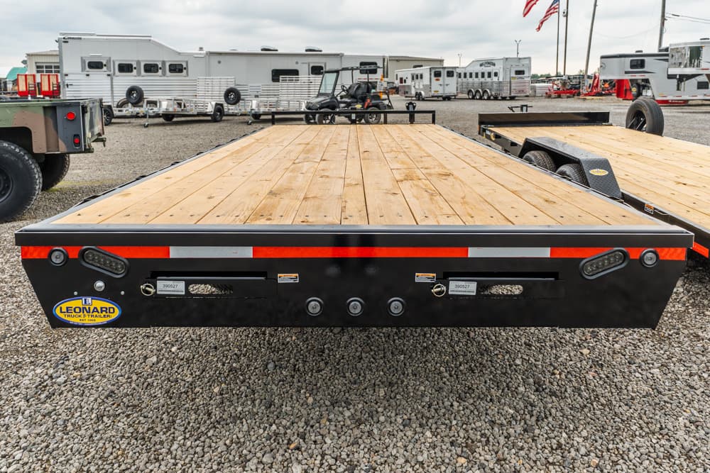 Flatbed trailer with a wooden deck and black frame, red/white reflective stripe, taillights, and a Leonard trailer sticker in a gravel lot.